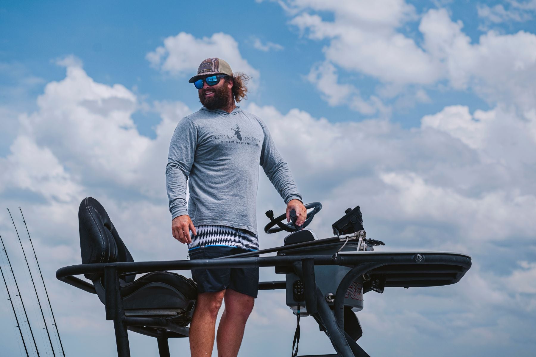 Captain Colt Brown wearing a gray long-sleeve Deer MEat for Dinner shirt, dark shorts, sunglasses, and a DMFD Life Mossy Oak bottomlands patch cap stands on a boat deck behind a console table, with a blue sky and white clouds in the background.