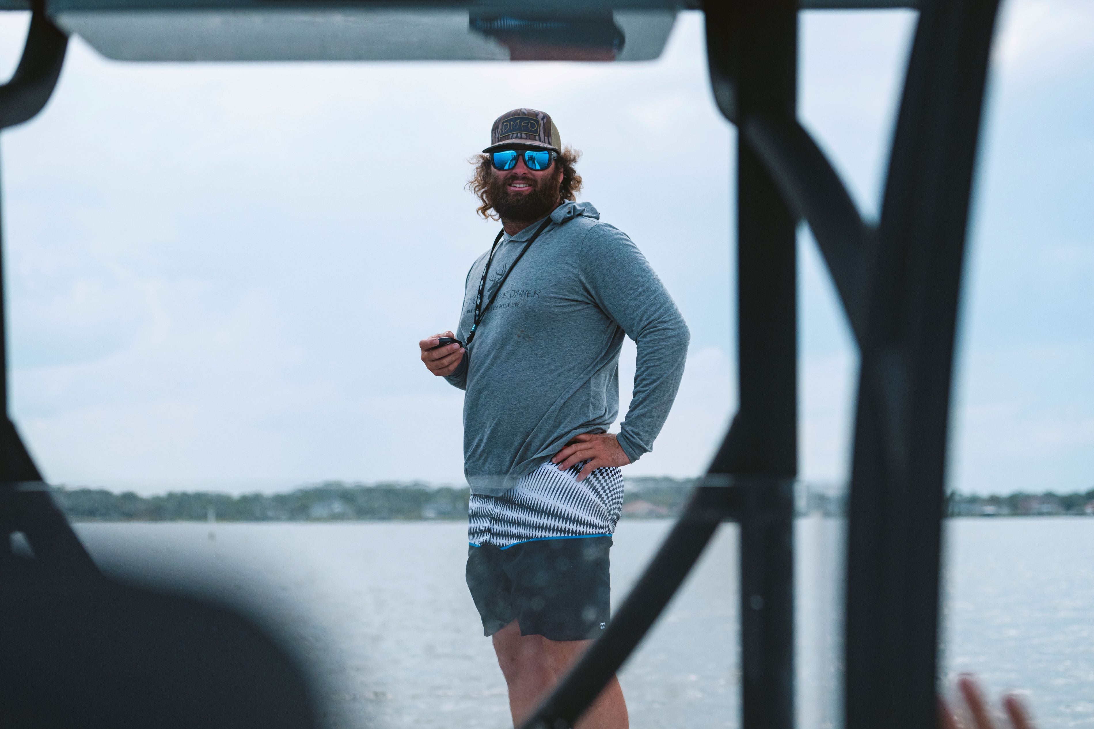 Captain Colt Brown wearing a gray Deer MEat for Dinner long-sleeve performance hoodie, dark swim trunks, a black cap, and blue mirrored sunglasses stands on a boat deck with water and a distant shoreline in the background using the Powerpole Move control on a lanyard