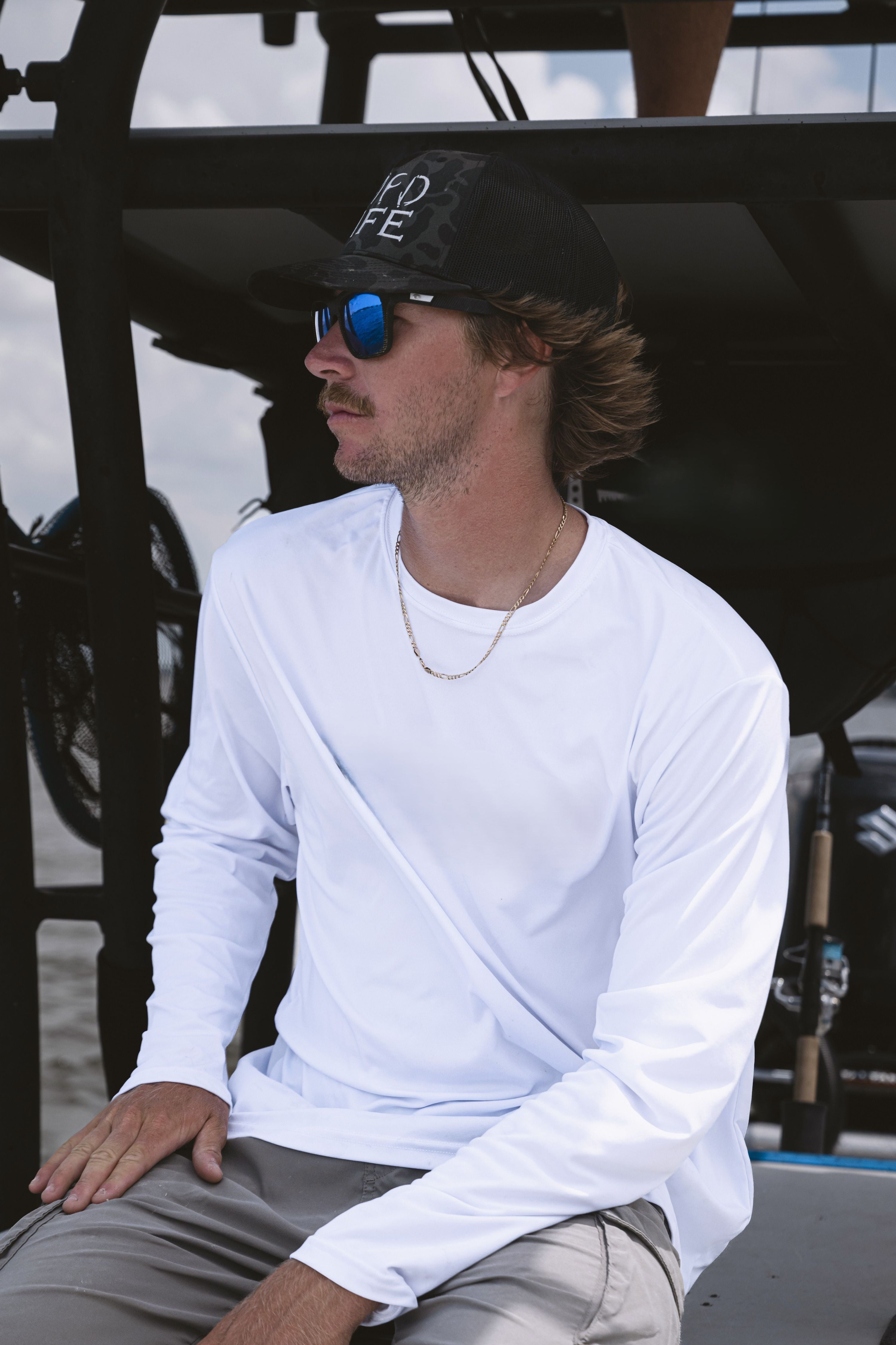 Captain Liam Garvey wearing a white long-sleeve crewneck shirt, gray pants, a black baseball cap with white lettering, and blue mirrored sunglasses, sitting on a boat.