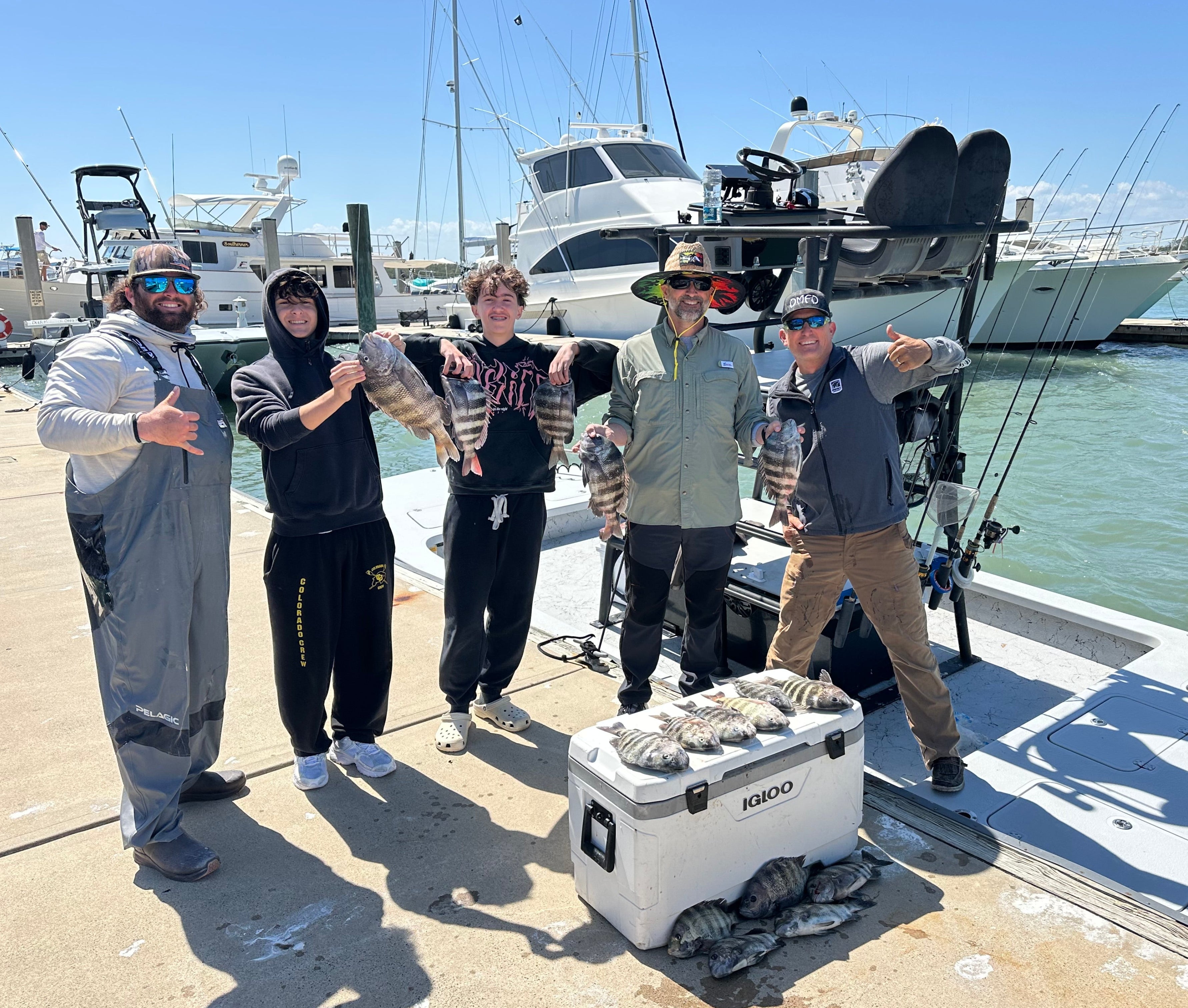 Group of five people on a boat with a cooler and fishing gear, docked at a marina.